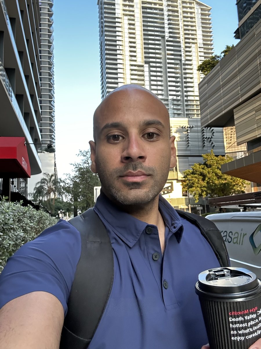 Malek Damiri in a navy polo with a backpack and a coffee, between downtown high-rises